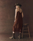 A woman faces away wearing The Legacy Dress by The Great Inc., styled with brown boots and a wide-brimmed hat, standing beside a wooden stool against a plain brown backdrop.