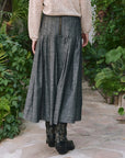 A person stands outdoors among greenery, wearing The Rhythm Skirt by The Great Inc., paired with black floral boots and a cream lace-texture long-sleeve top.