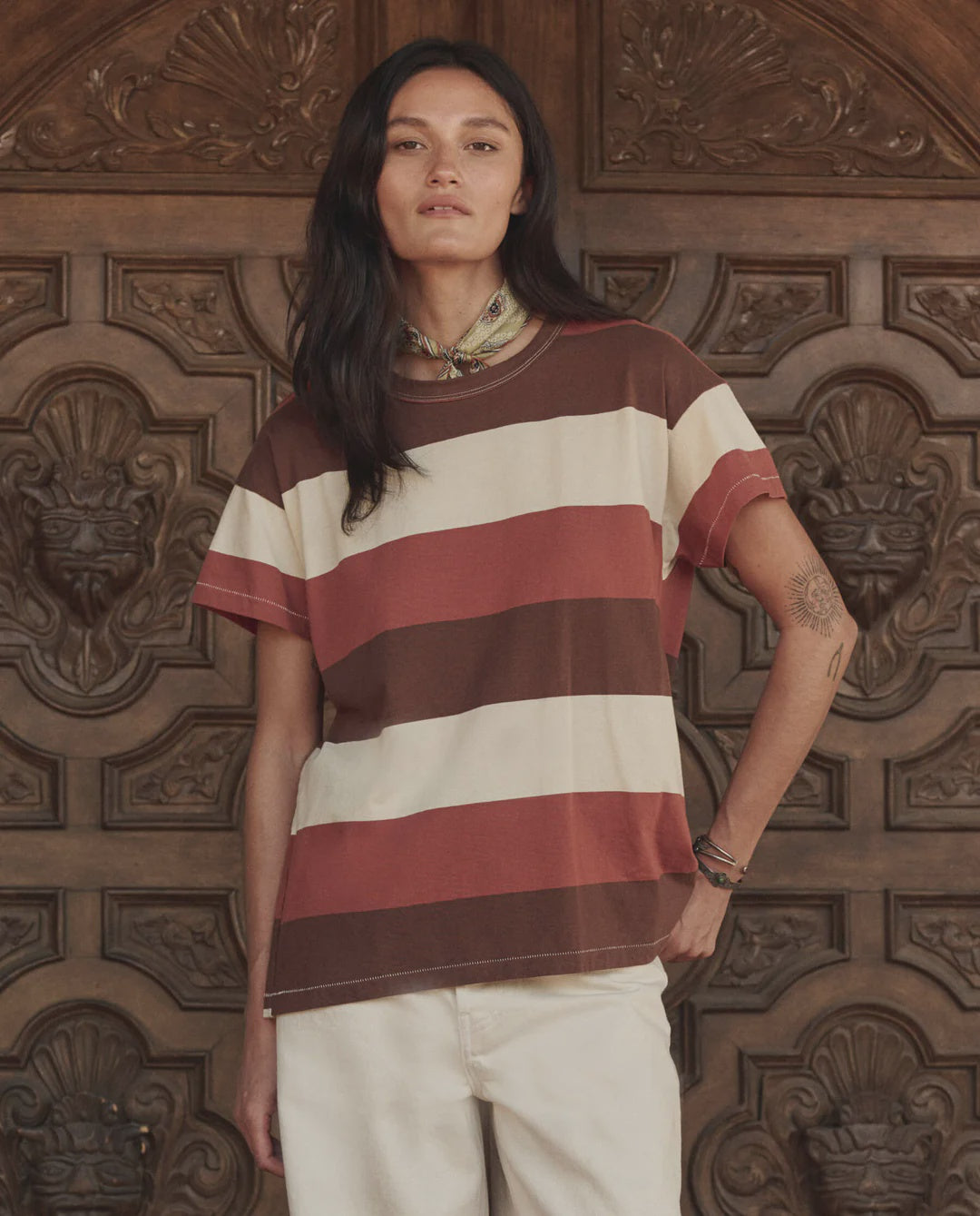 A person with long dark hair wearing The Boxy Crew 26 tee by The Great Inc. in brown, white, and red stripes, light pants, and a bandana stands before an ornate carved wooden door.