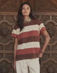 A person with long dark hair wearing The Boxy Crew 26 tee by The Great Inc. in brown, white, and red stripes, light pants, and a bandana stands before an ornate carved wooden door.