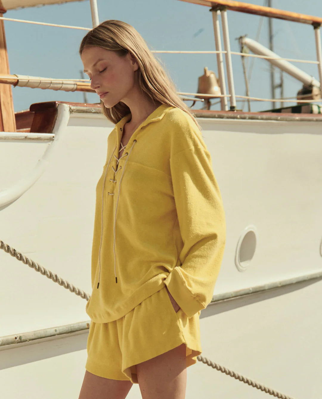 Wearing The Great Inc.'s The Micro Terry Swing Short, a woman in yellow stands before a white boat on a sunny day, hands in her pockets, looking down.