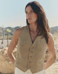 A person with long dark hair stands outdoors in a desert landscape, wearing The Great Inc.'s The Suit Vest in light brown cotton herringbone paired with white pants, mountains and cacti visible in the background.