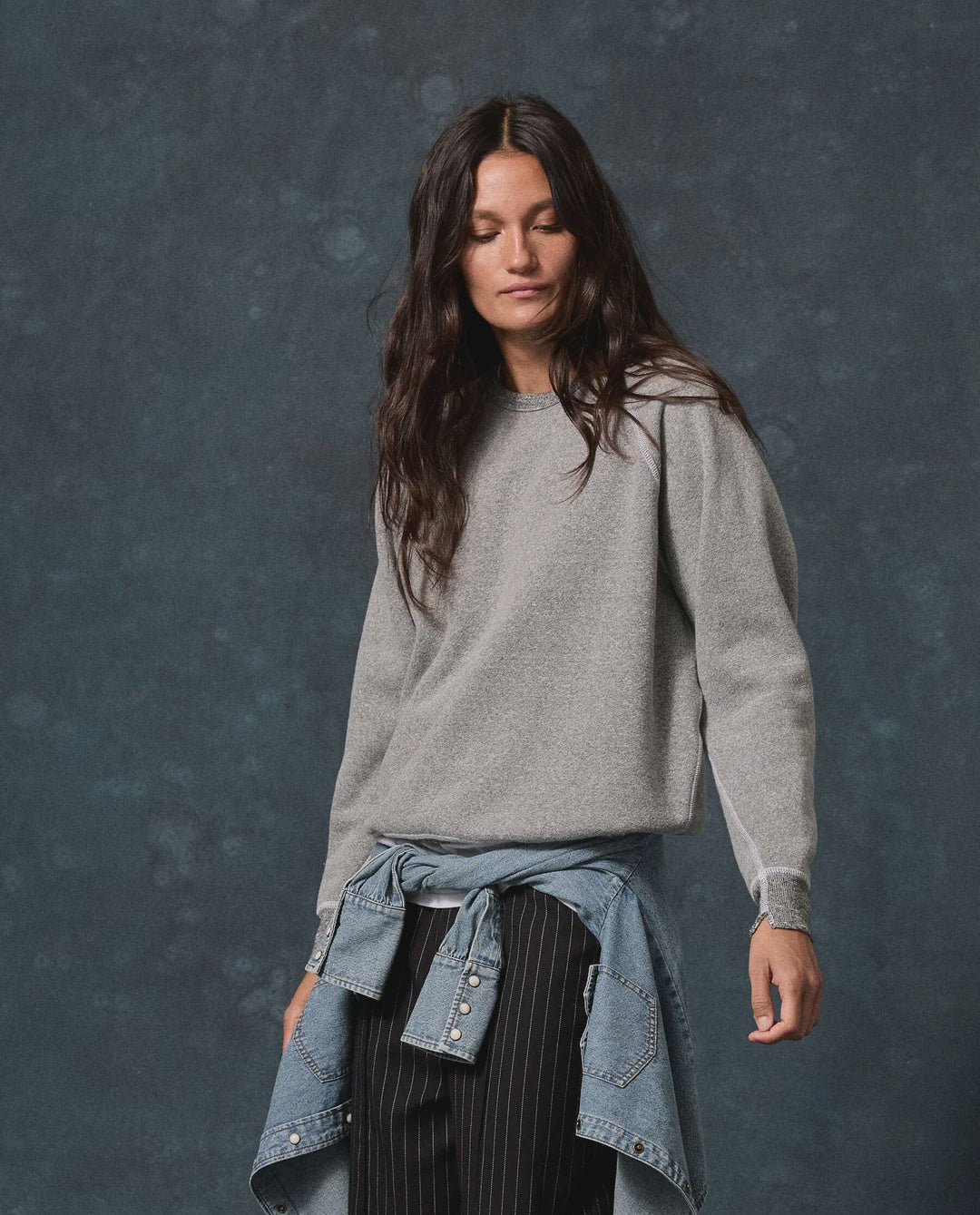The Great Inc. fleece college sweatshirt styled with a denim shirt at the waist, shown against a dark background.