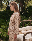 A woman in The Great Inc.’s The Pajama Shirt stands in profile outdoors beside a bed with a teddy bear, surrounded by lush green plants and trees.