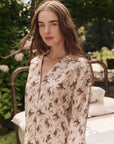 A woman with long brown hair stands outdoors, wearing The Pajama Shirt by The Great Inc., a light-colored blouse inspired by vintage pajama tops. Green foliage and part of a metal bed frame are visible in the background as she looks at the camera.