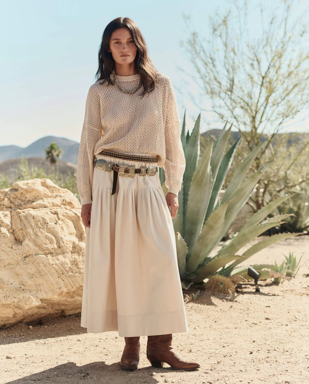 A woman stands outdoors in a desert landscape, wearing The Highland Skirt by The Great Inc., a knit sweater, brown boots, and a pouch belt. Large agave plants and rocky terrain under a clear sky complete the vintage look.