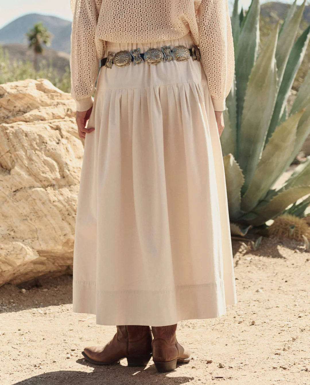 A person stands outdoors on sandy ground wearing The Great Inc.'s cream-colored Highland Skirt, a loose knit sweater, brown cowboy boots, and a silver belt, with desert plants and rocks in the background.