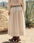 A person stands outdoors on sandy ground wearing The Great Inc.'s cream-colored Highland Skirt, a loose knit sweater, brown cowboy boots, and a silver belt, with desert plants and rocks in the background.