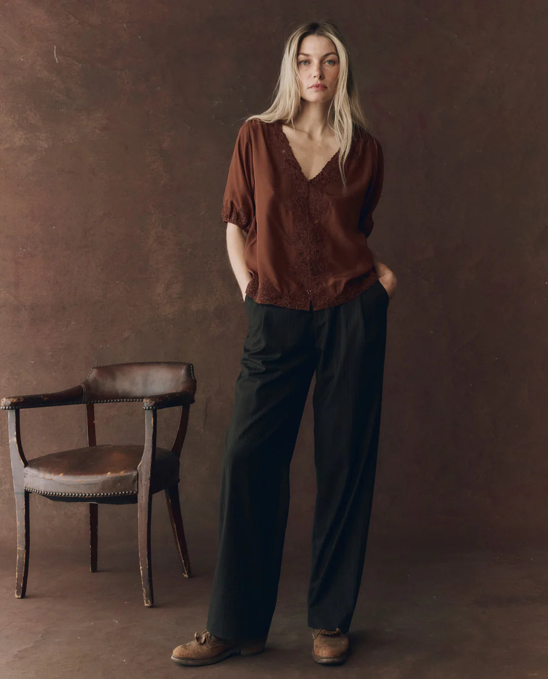 A woman with long blonde hair stands by a vintage wooden chair, wearing The SS Legacy Top by The Great Inc., paired with loose black trousers and brown shoes against a brown textured backdrop.