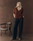 A woman with long blonde hair stands by a vintage wooden chair, wearing The SS Legacy Top by The Great Inc., paired with loose black trousers and brown shoes against a brown textured backdrop.