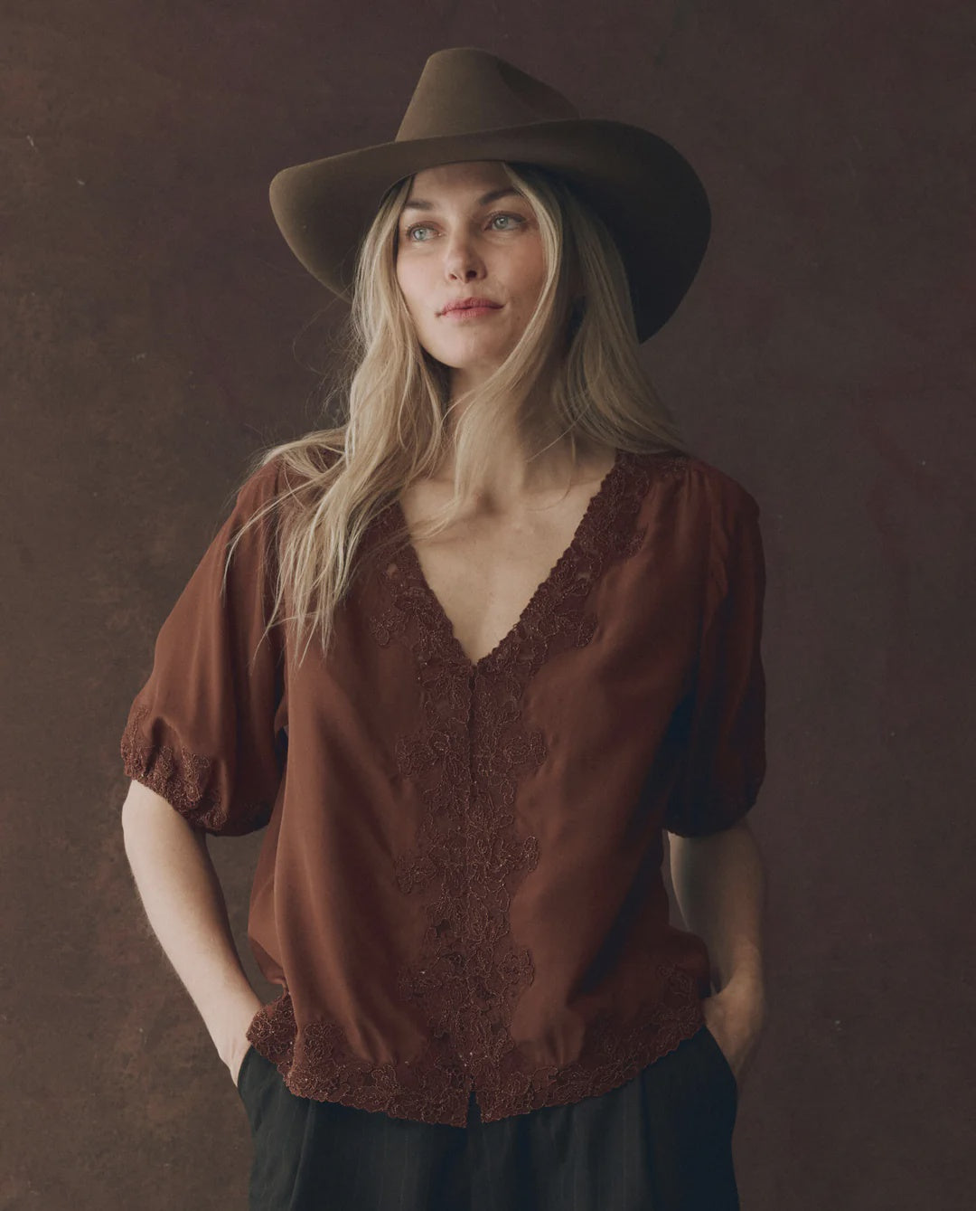 A woman with long blonde hair wears The SS Legacy Top by The Great Inc., a brown silk V-neck top with embroidered mesh lace, paired with a brown wide-brimmed hat and standing against a dark textured background.