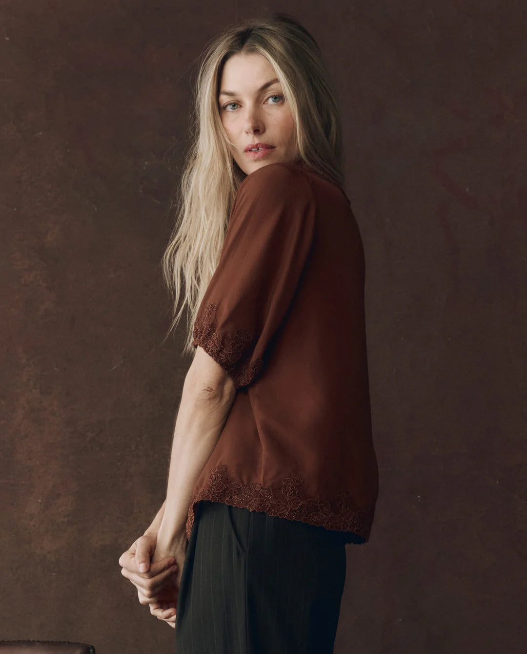 A woman with long blonde hair models The Great Inc.'s SS Legacy Top—a brown embroidered mesh lace blouse—paired with black pants, standing sideways and looking at the camera against a dark brown textured backdrop.