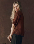 A woman with long blonde hair models The Great Inc.'s SS Legacy Top—a brown embroidered mesh lace blouse—paired with black pants, standing sideways and looking at the camera against a dark brown textured backdrop.