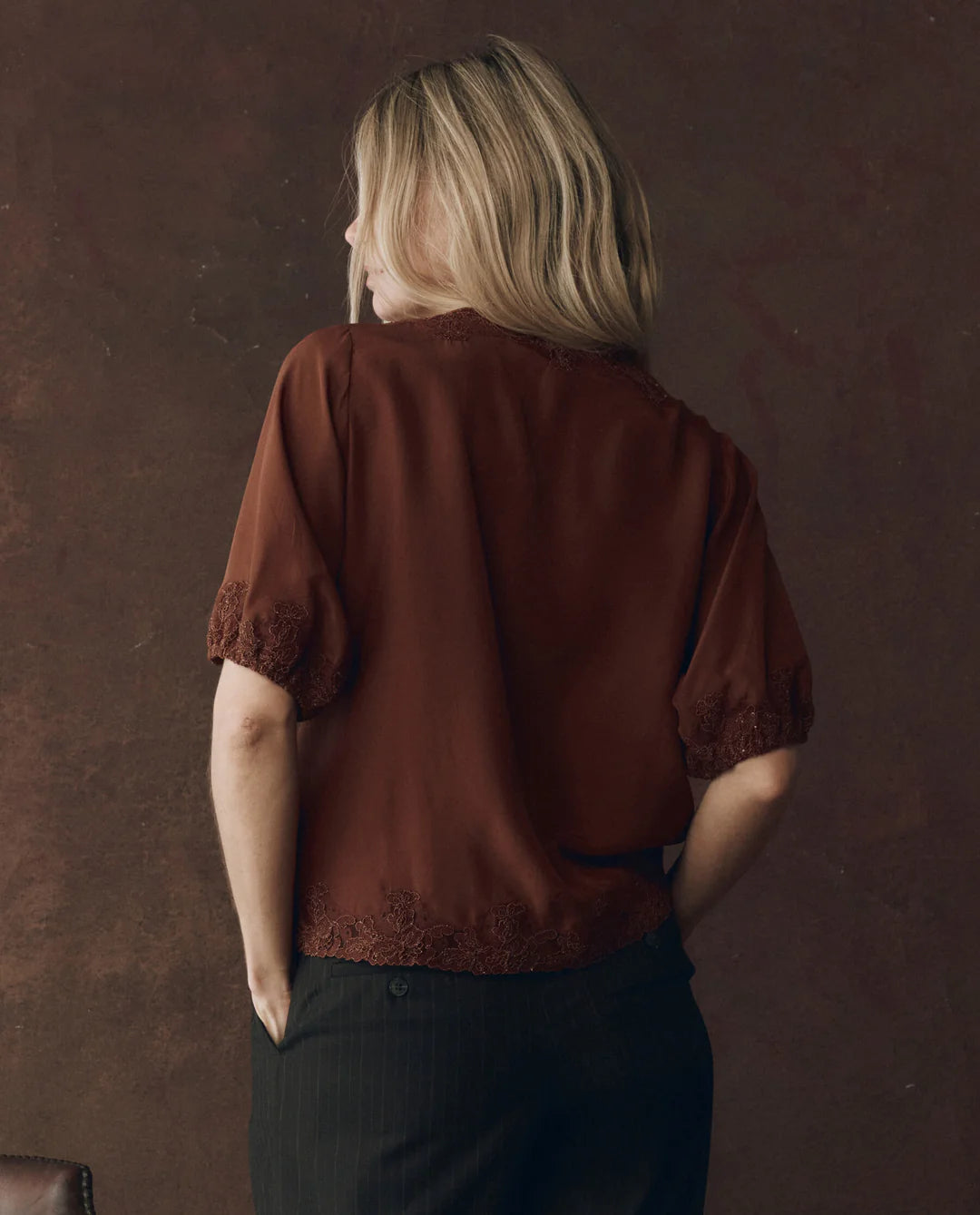 A person with blonde hair, seen from behind, wears The SS Legacy Top by The Great Inc.—a short-sleeved, dark red silk blouse with embroidered mesh lace trim—paired with black pinstripe pants against a 1920s loungewear-inspired backdrop.