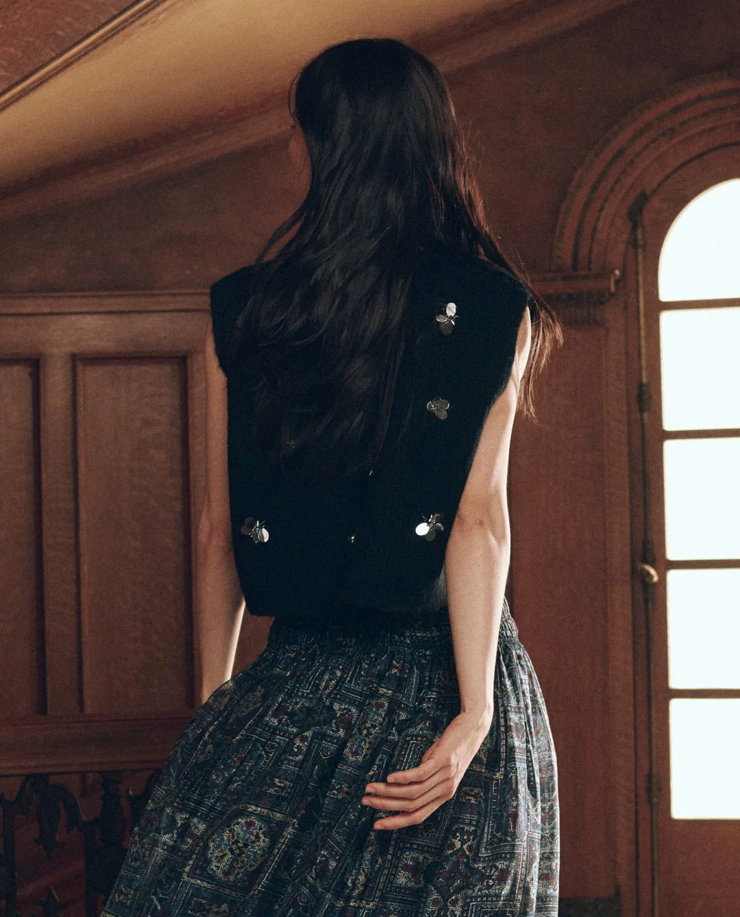 Black embellished sweater vest with silver bee accents, styled over a patterned skirt.