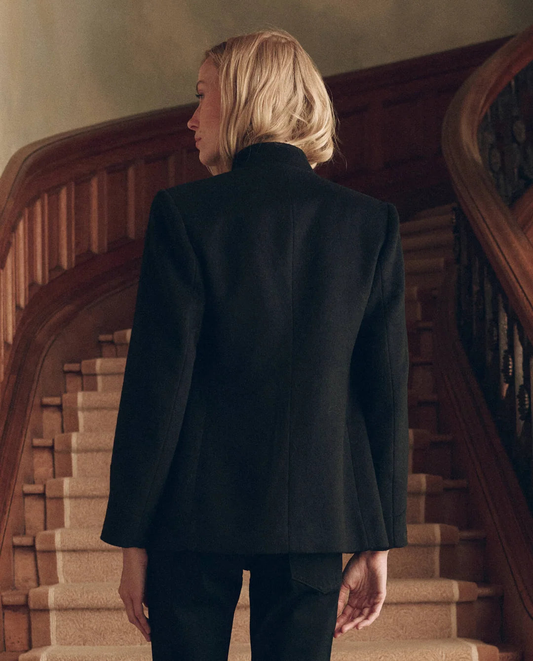 Vintage The Military Blazer in black wool worn by a woman standing before a curved wooden staircase.
