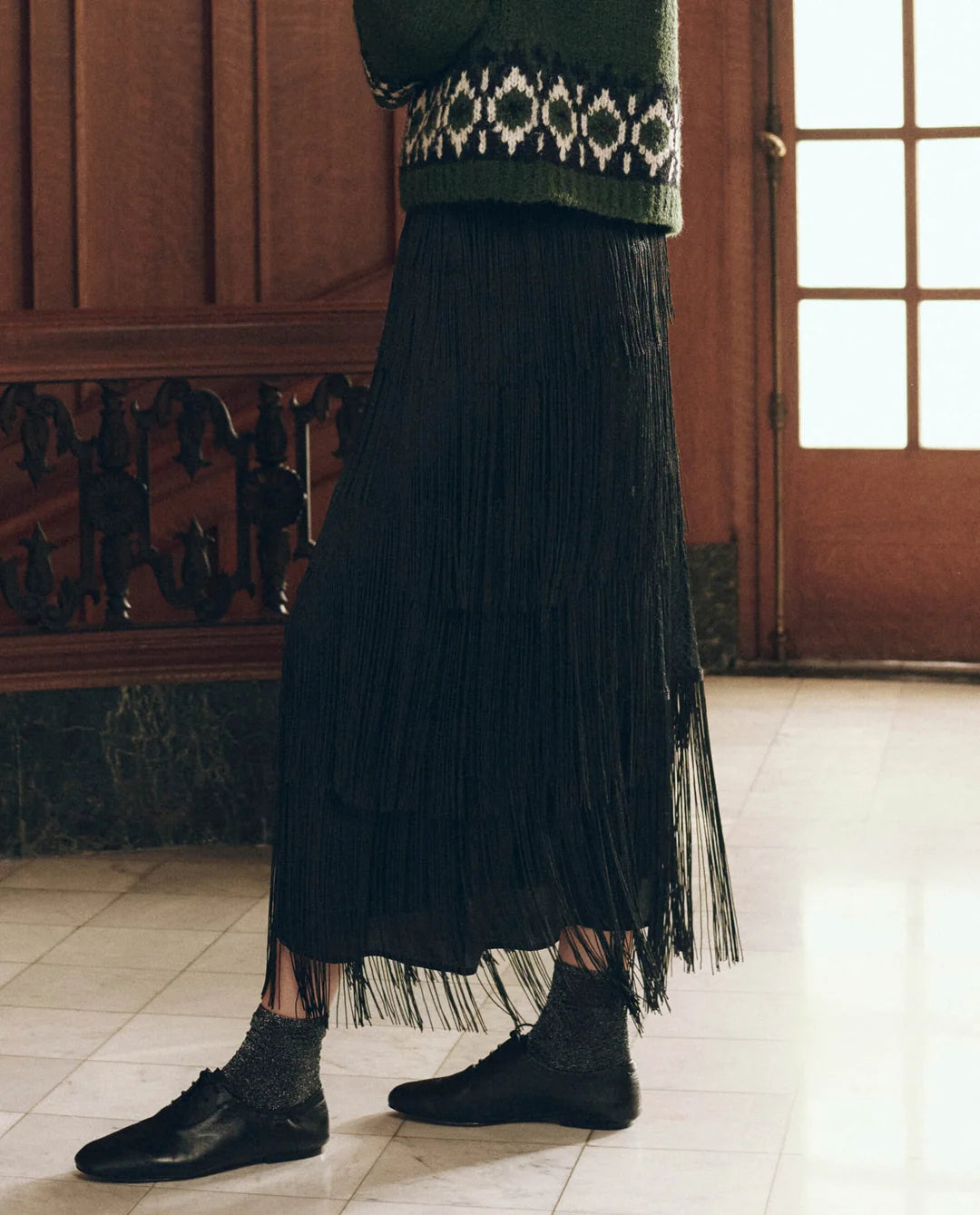 The Fringe Skirt in black tiered fringe styled with a green patterned sweater, dark socks, and black shoes.