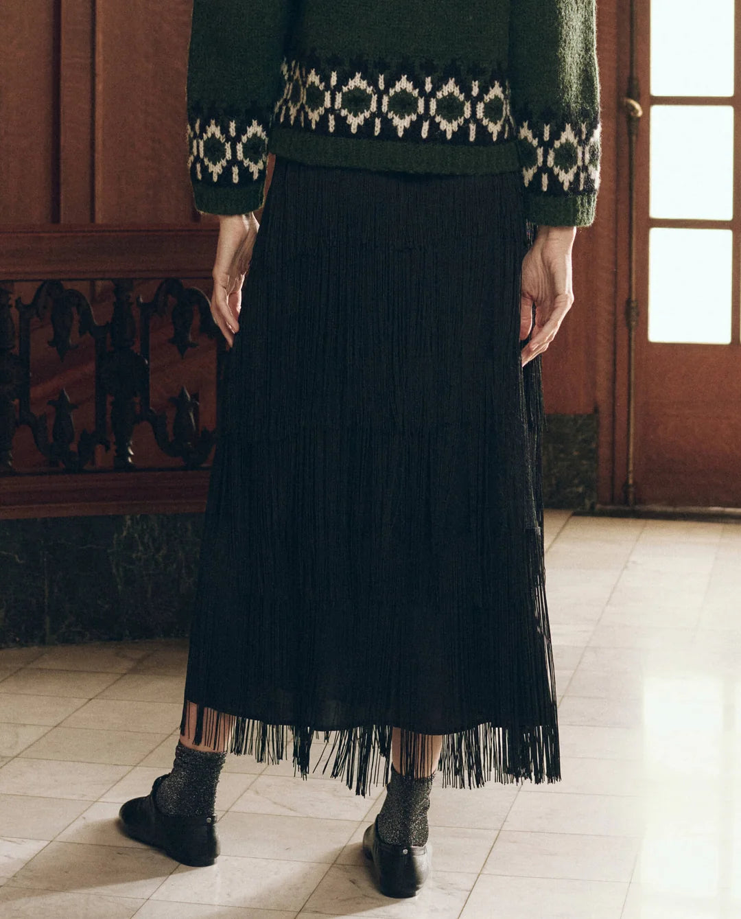 Black midi fringe skirt by The Great Inc. paired with a green sweater and dark shoes in a 1920s-inspired style.