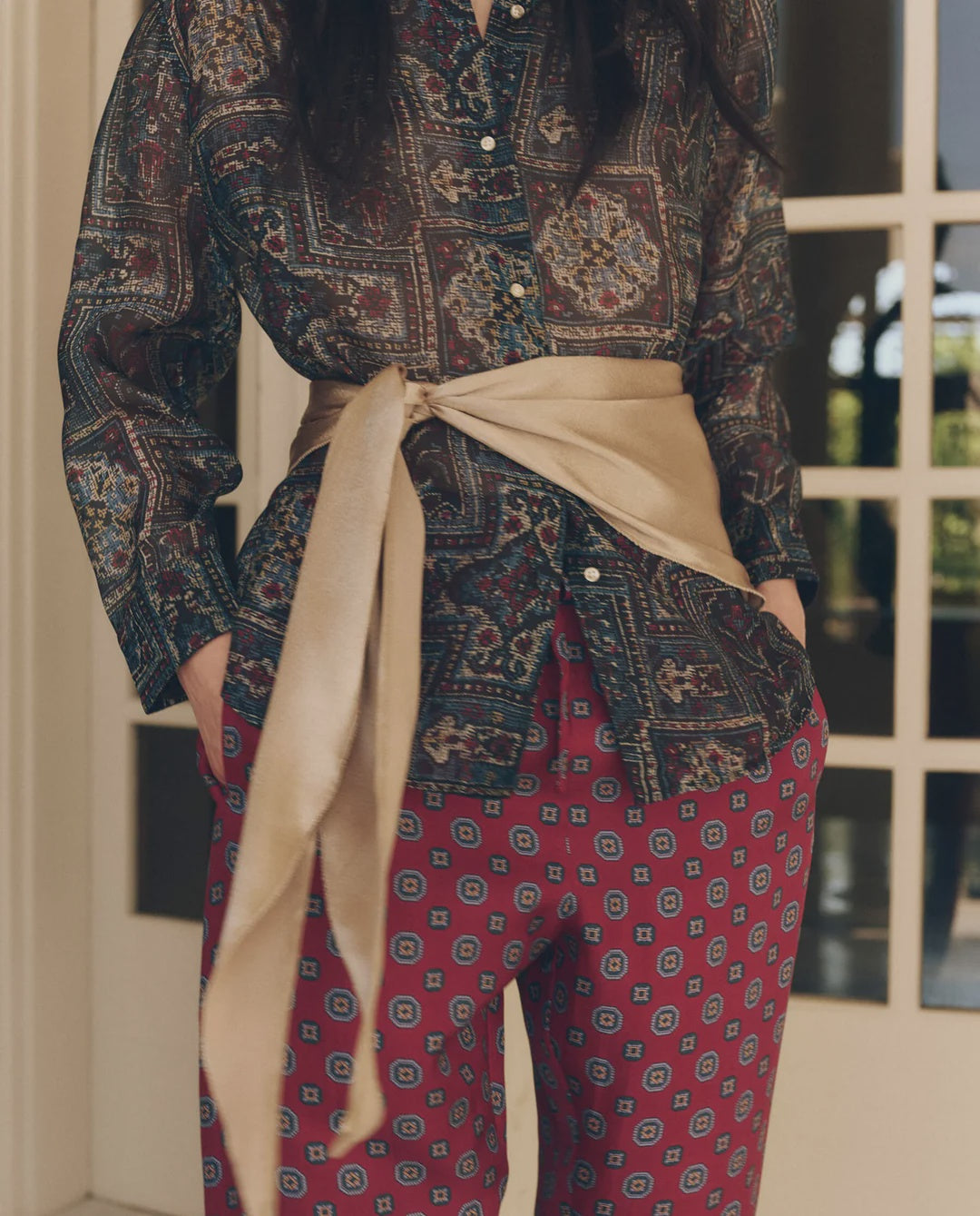 Beige The Wrap Scarf tied at the waist over a patterned button-down and red geometric trousers.