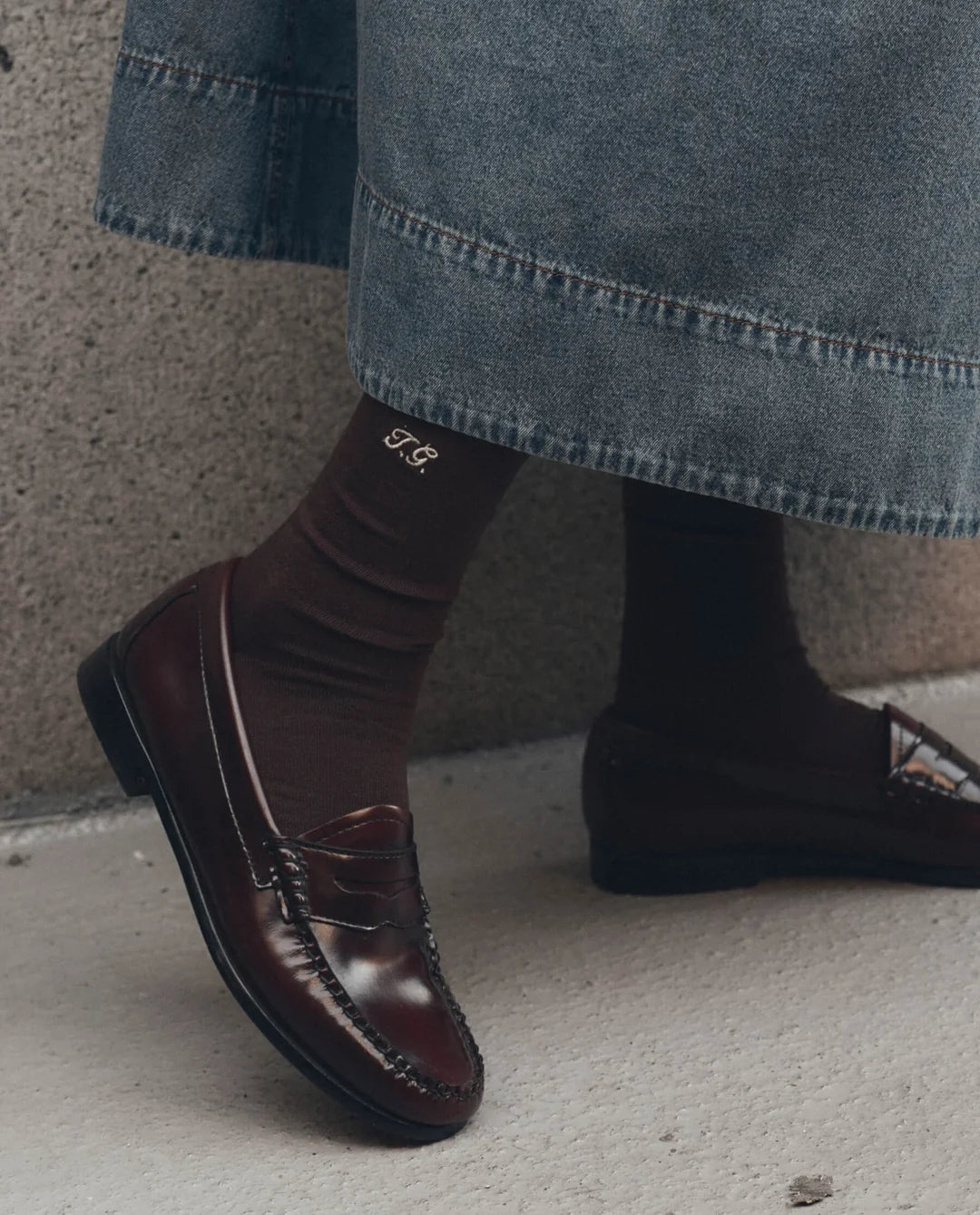 The Great Inc. Monogrammed Trouser Sock in brown, styled with loafers and a denim skirt on concrete.