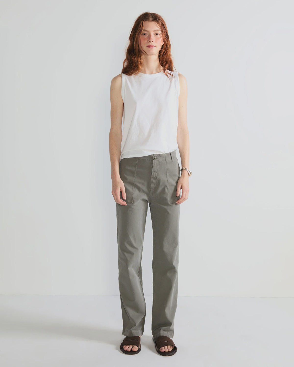 A person with long reddish hair wears Kule’s The Tank, paired with gray straight-leg pants and black sandals, standing against a plain white background.