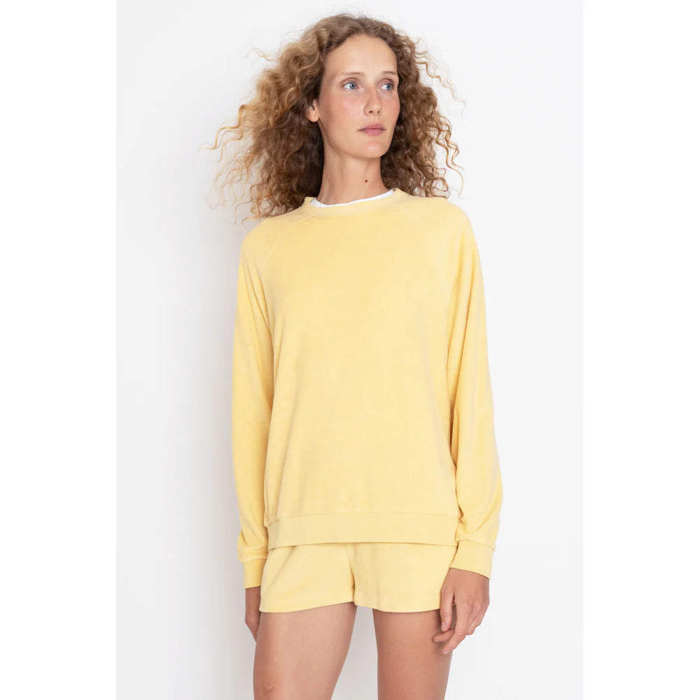 A person with curly hair wears The Terry Franny sweatshirt by Kule in light yellow, paired with matching shorts. The plain white background enhances the scene as they gaze slightly to the side.