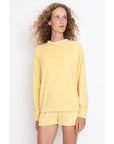 A person with curly hair wears The Terry Franny sweatshirt by Kule in light yellow, paired with matching shorts. The plain white background enhances the scene as they gaze slightly to the side.