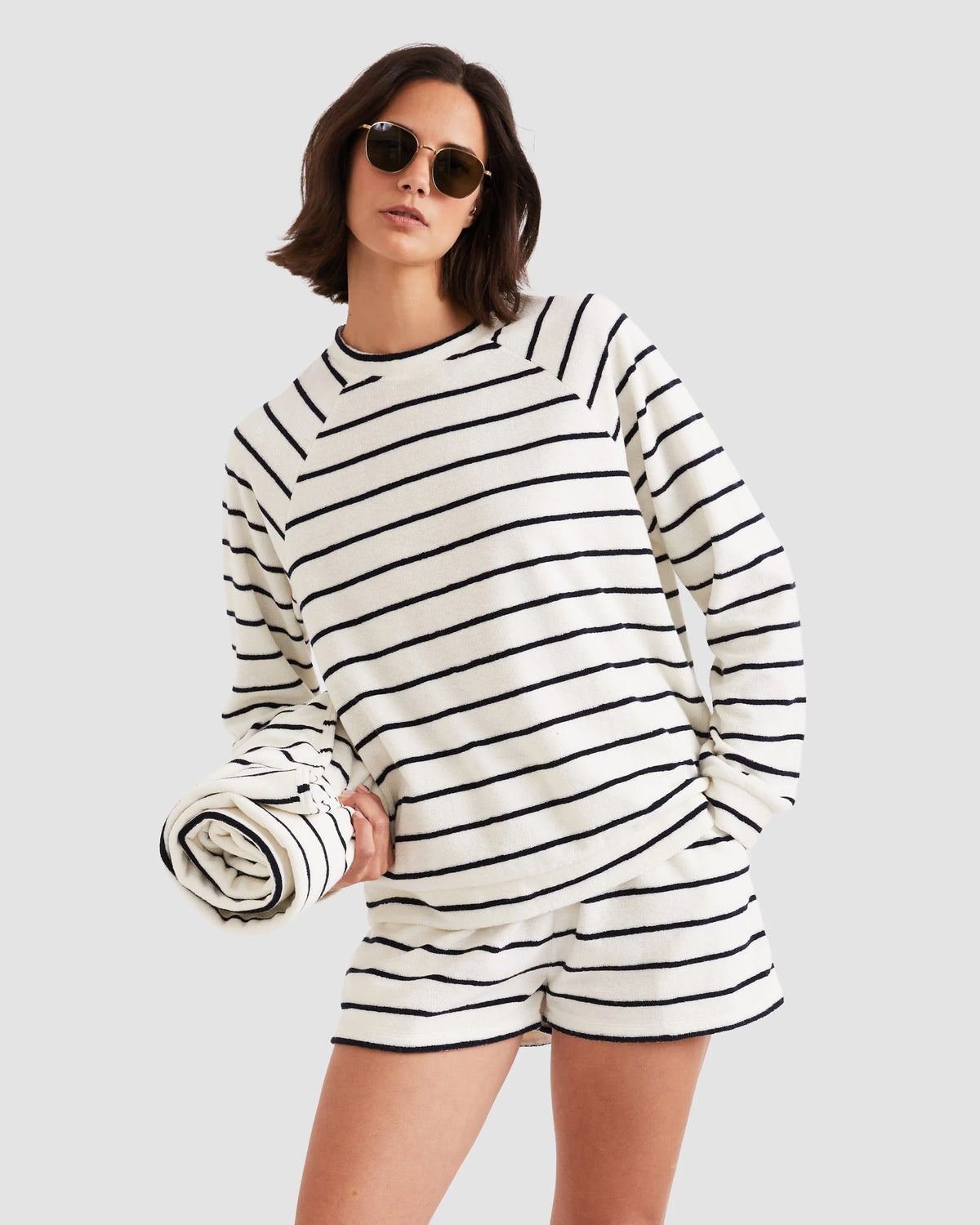 A woman wearing the Kule Terry Franny sweatshirt with matching striped shorts and sunglasses holds a rolled-up striped towel against a plain light gray background.