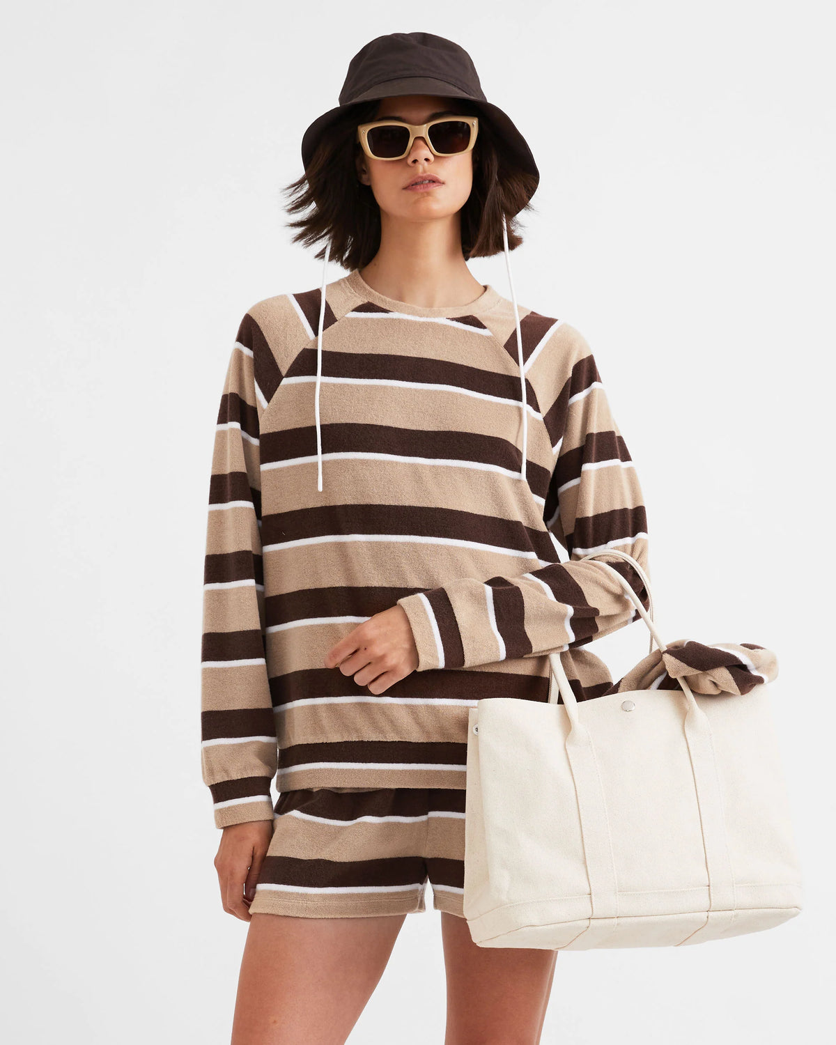 A woman wearing The Terry Franny sweatshirt by Kule, paired with matching shorts, a black bucket hat, sunglasses, and holding a large cream canvas tote against a plain background.