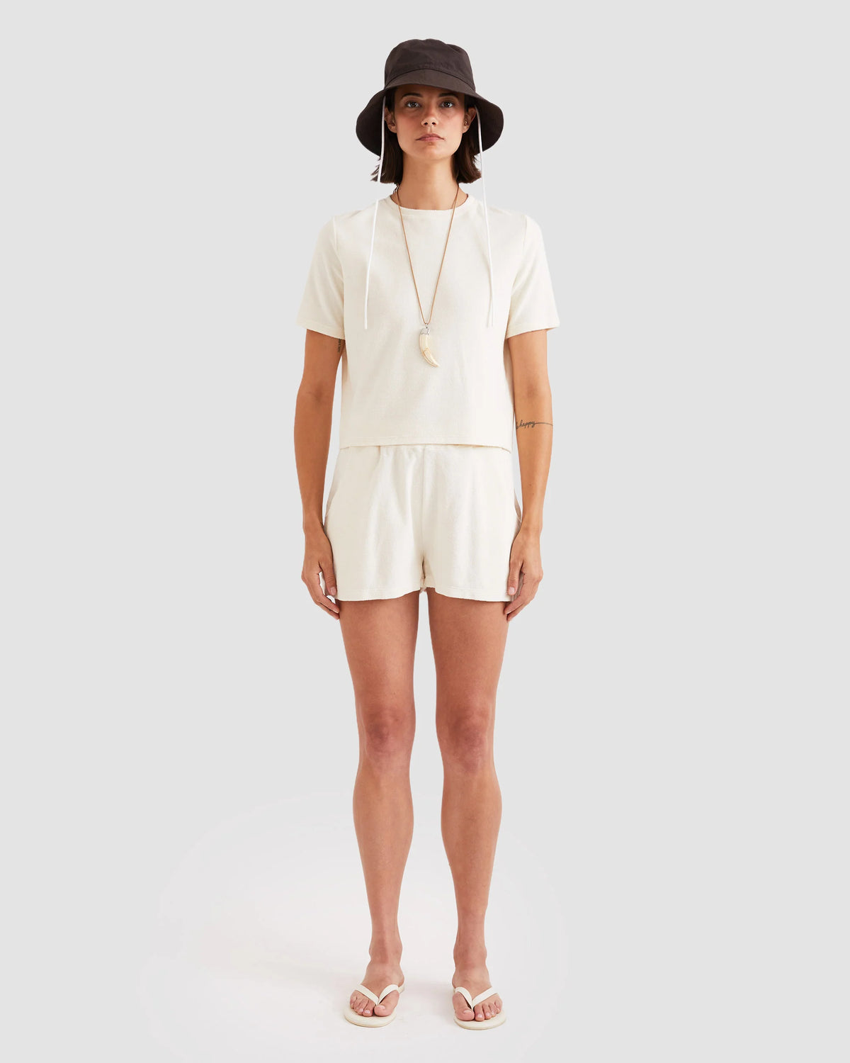 A woman stands against a plain background, wearing Kule's The Terry Hurley cream cotton-blend cropped tee and shorts set, paired with a black bucket hat, white sandals, and a long necklace.