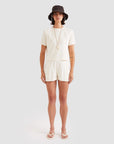 A woman stands against a plain background, wearing Kule's The Terry Hurley cream cotton-blend cropped tee and shorts set, paired with a black bucket hat, white sandals, and a long necklace.