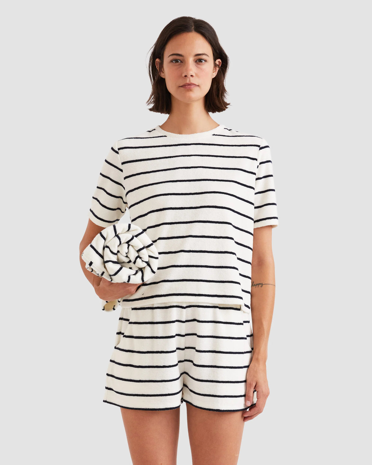 A woman wears Kule’s The Terry Hurley—matching white cotton-blend cropped tee and shorts with black stripes—while holding a rolled-up striped towel against a plain light gray background.