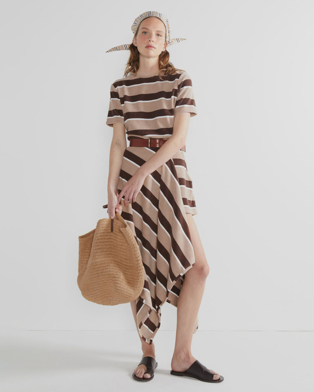 A woman stands against a white background wearing The Terry Hurley dress by Kule, a brown belt, headscarf, black sandals, and holding a large woven tote bag.