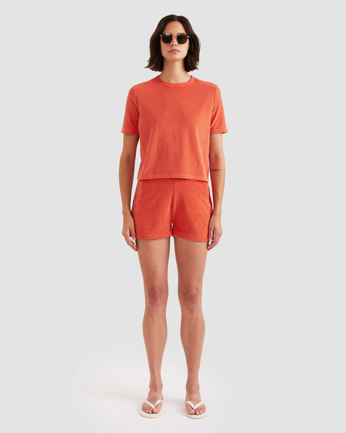 A person in Kule's The Terry Hurley—a roomy cotton-blend cropped tee and shorts set—wears sunglasses and white flip-flops, standing against a plain light gray background.