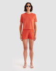 A person in Kule's The Terry Hurley—a roomy cotton-blend cropped tee and shorts set—wears sunglasses and white flip-flops, standing against a plain light gray background.