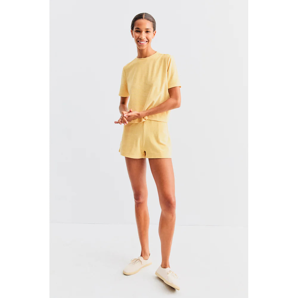 A person stands smiling in a minimalist, light-filled space, wearing Kule's matching cotton-blend yellow T-shirt and The Terry Venus short with an elastic waist, paired with cream-colored sneakers.