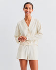 A smiling person with dark hair tied back wears Kule’s cream-colored The Terry Venus short and a matching cardigan, standing against a plain light background.