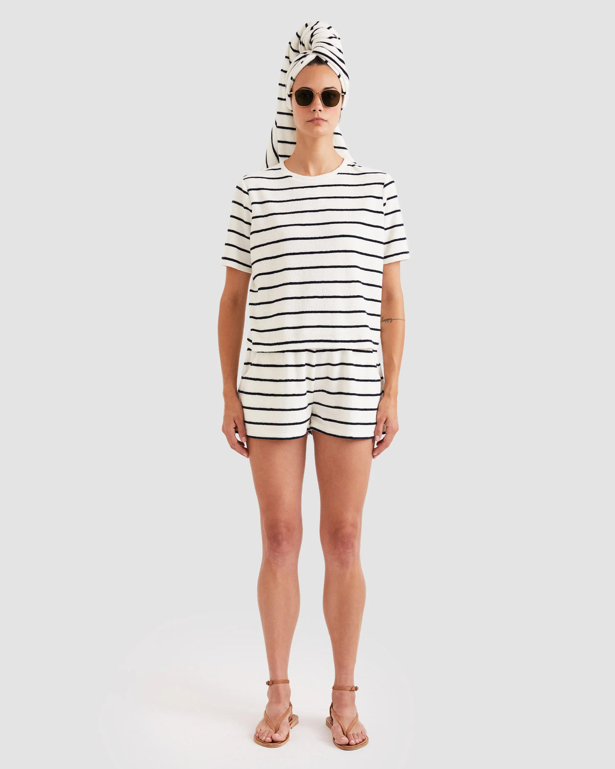 A person stands facing forward against a plain background, wearing sunglasses, a white striped T-shirt, and Kule's The Terry Venus short—a cotton-blend style with an elastic waist. A towel is wrapped around their head; they are barefoot and neutral-faced.