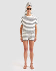A person stands facing forward against a plain background, wearing sunglasses, a white striped T-shirt, and Kule's The Terry Venus short—a cotton-blend style with an elastic waist. A towel is wrapped around their head; they are barefoot and neutral-faced.
