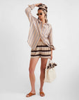 A woman poses in a striped shirt, Kule The Terry Venus shorts, brown sandals, and sunglasses. She carries a tote bag and has a matching striped scarf or towel wrapped around her head.