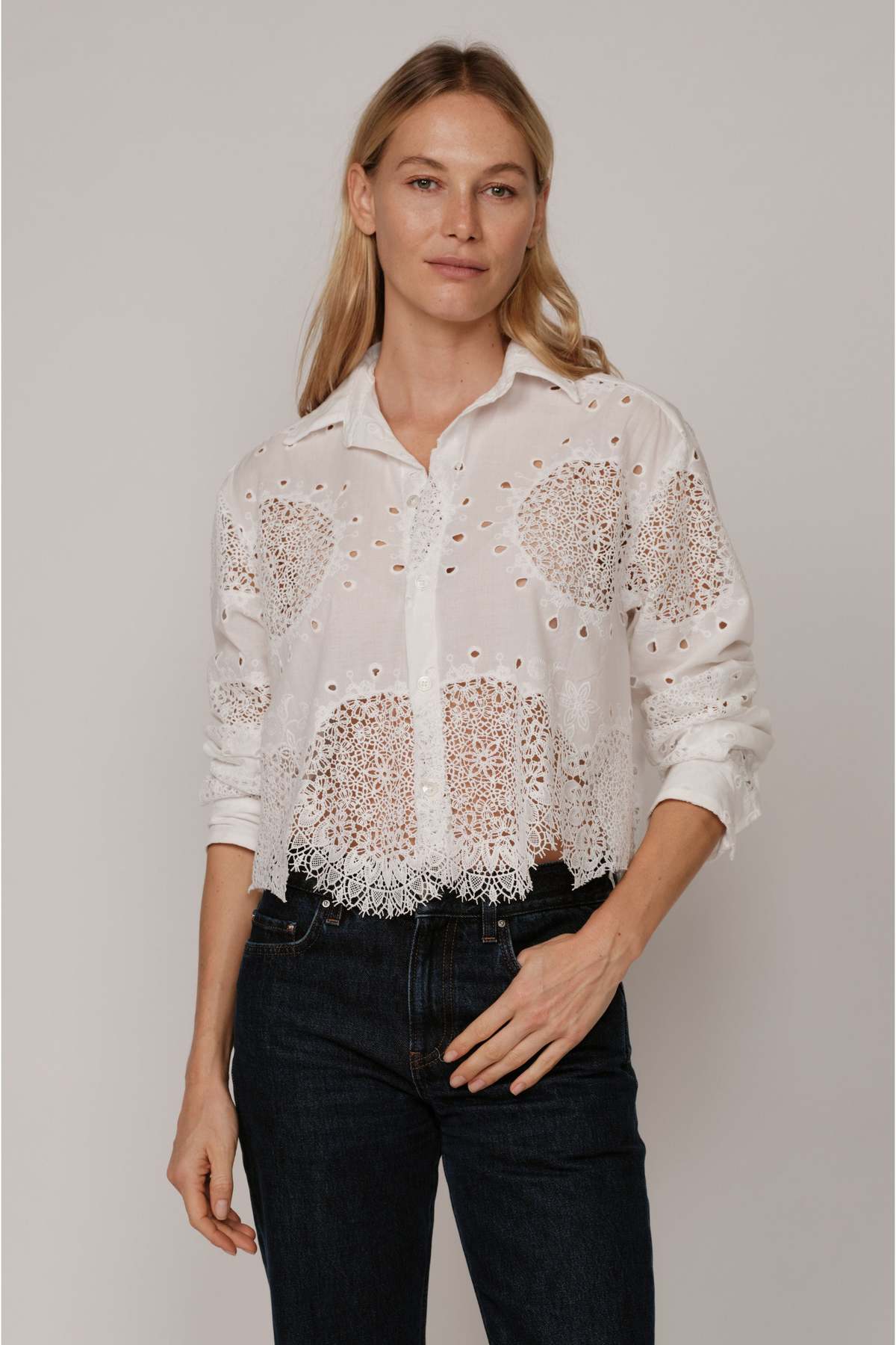 A woman with long blonde hair wears The Belle by Cissa—a white, lace-patterned, relaxed fit shirt with a fringe hem—paired with dark jeans, standing against a plain light background.