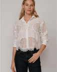 A woman with long blonde hair wears The Belle by Cissa—a white, lace-patterned, relaxed fit shirt with a fringe hem—paired with dark jeans, standing against a plain light background.
