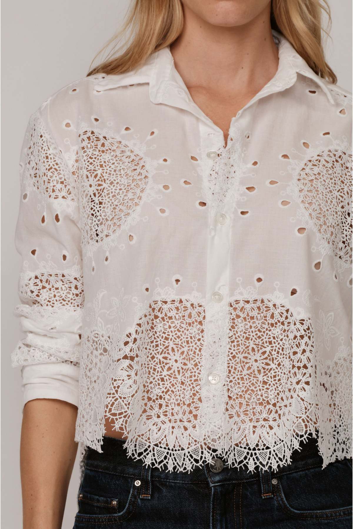 A woman wears The Belle shirt by Cissa—a white, long-sleeve, button-up Egyptian cotton top with intricate lace cut-out details and a scalloped hem—paired with dark denim jeans for an effortlessly elegant look.