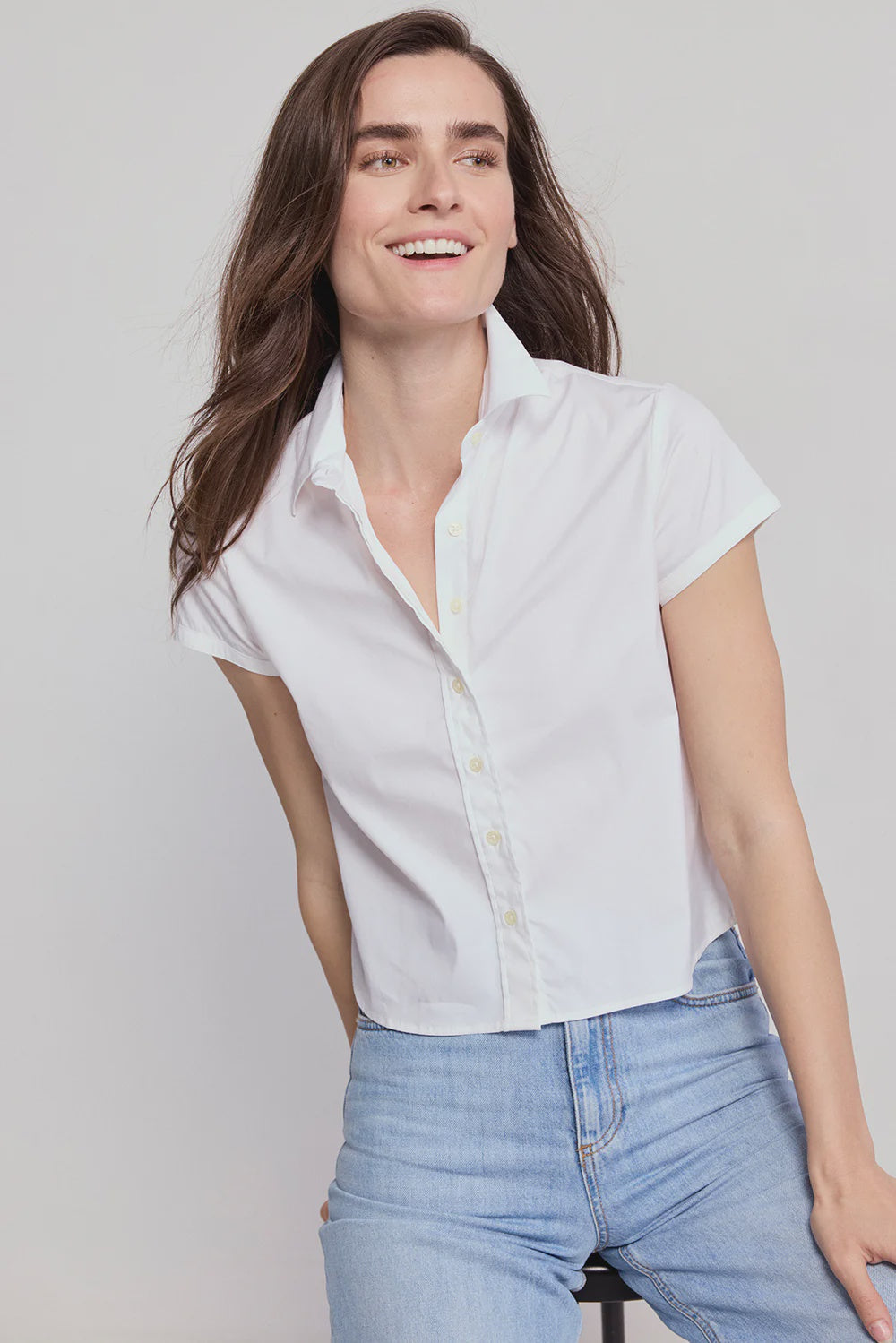 A woman with long brown hair wears THE SHIRT’s Boxy Short Sleeve Cropped Shirt in white and light blue jeans, smiling as she leans to the side against a plain light background.