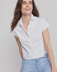 A woman with long brown hair wears THE SHIRT’s Boxy Short Sleeve Cropped Shirt in white and light blue jeans, smiling as she leans to the side against a plain light background.