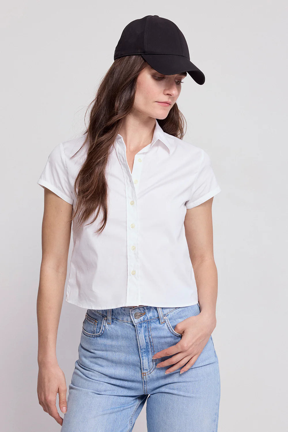 A person wears a black baseball cap, light blue jeans, and THE SHIRT’s The Boxy Short Sleeve Cropped Shirt with No Gape button technology, standing against a plain light background and looking down with one hand in their pocket.
