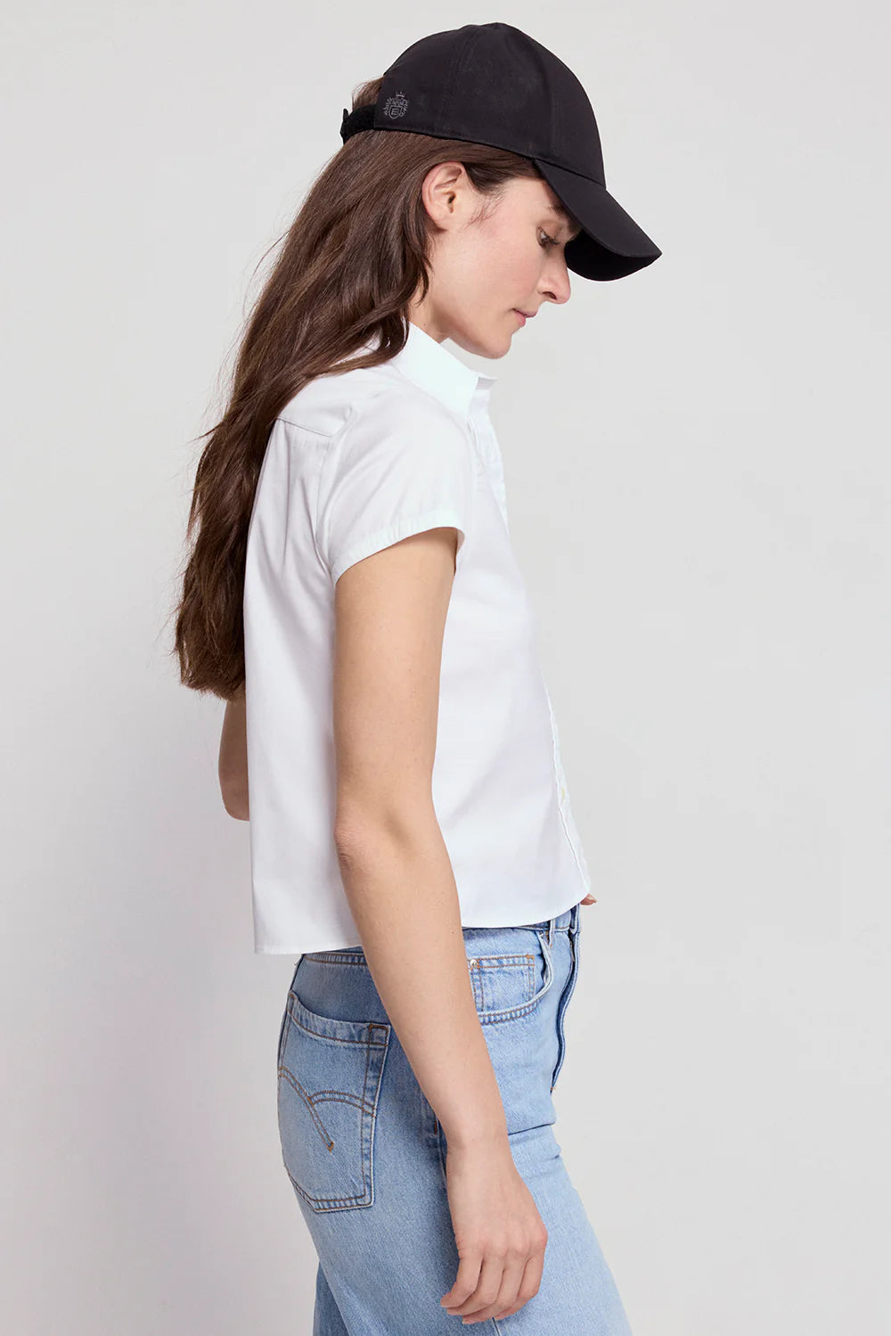 A person with long brown hair wears THE SHIRT's The Boxy Short Sleeve Cropped Shirt, paired with a black baseball cap and light blue jeans, standing in side profile against a plain light background.