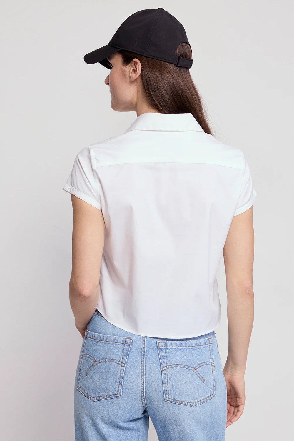 A woman with long brown hair is seen from the back wearing a black baseball cap and THE SHIRT’s Boxy Short Sleeve Cropped Shirt, featuring No Gape button technology, paired with light blue jeans against a plain light background.