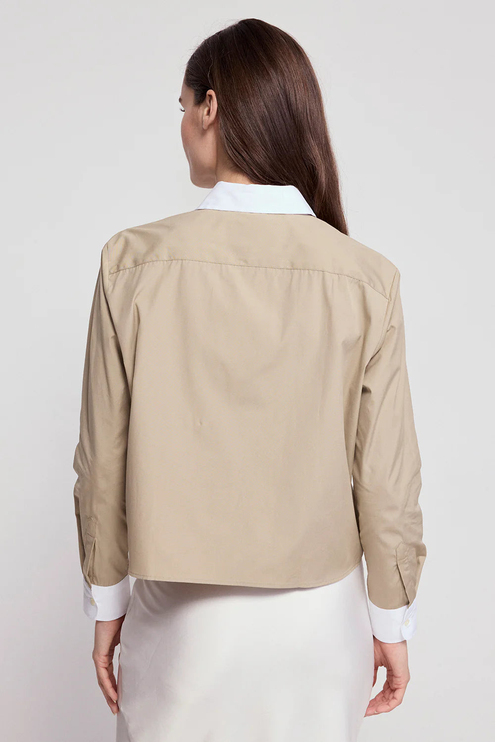 A woman with long brown hair poses with her back to the camera, wearing THE SHIRT's The LS Boxy Crop beige jacket with white collar and cuffs over a white skirt, against a plain light background.