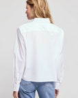 A blonde woman wearing THE SHIRT’s The LS Boxy Crop, a long-sleeve white button-up with a No Gape feature, and blue jeans stands facing away from the camera against a light gray background.